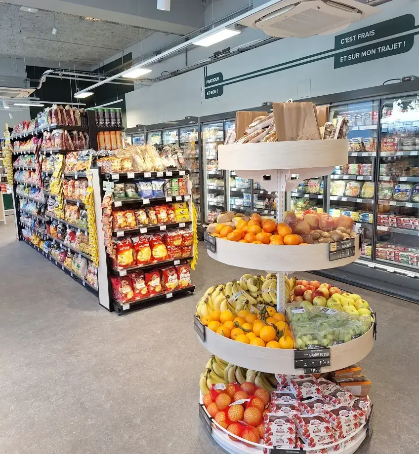 Intérieur du Carrefour Express en gare de Chambéry, présentoir de fruits frais au premier plan, rayonnages épicerie et réfrigérateurs en fond